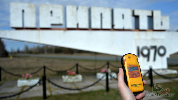 Рука с дозиметром на фоне стелы города Припять