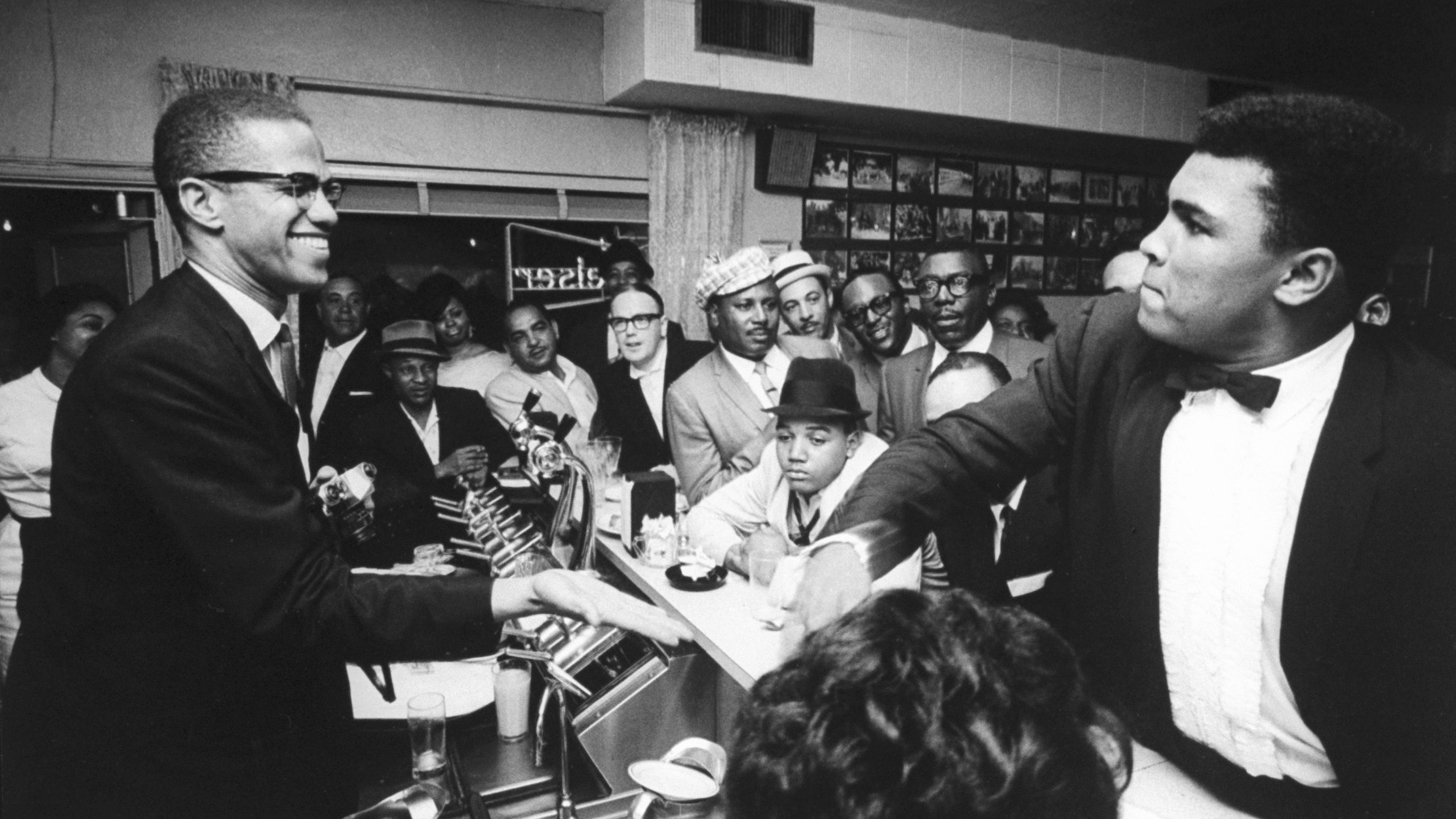 March 1964 - Miami: Black Muslim leader Malcolm X (L), behind soda fountain, with tux-clad Cassius Clay (now Muhammad Ali) (R), while surrounded by jubilant fans after he beat Sonny Liston for the heavyweight championship of the world. 