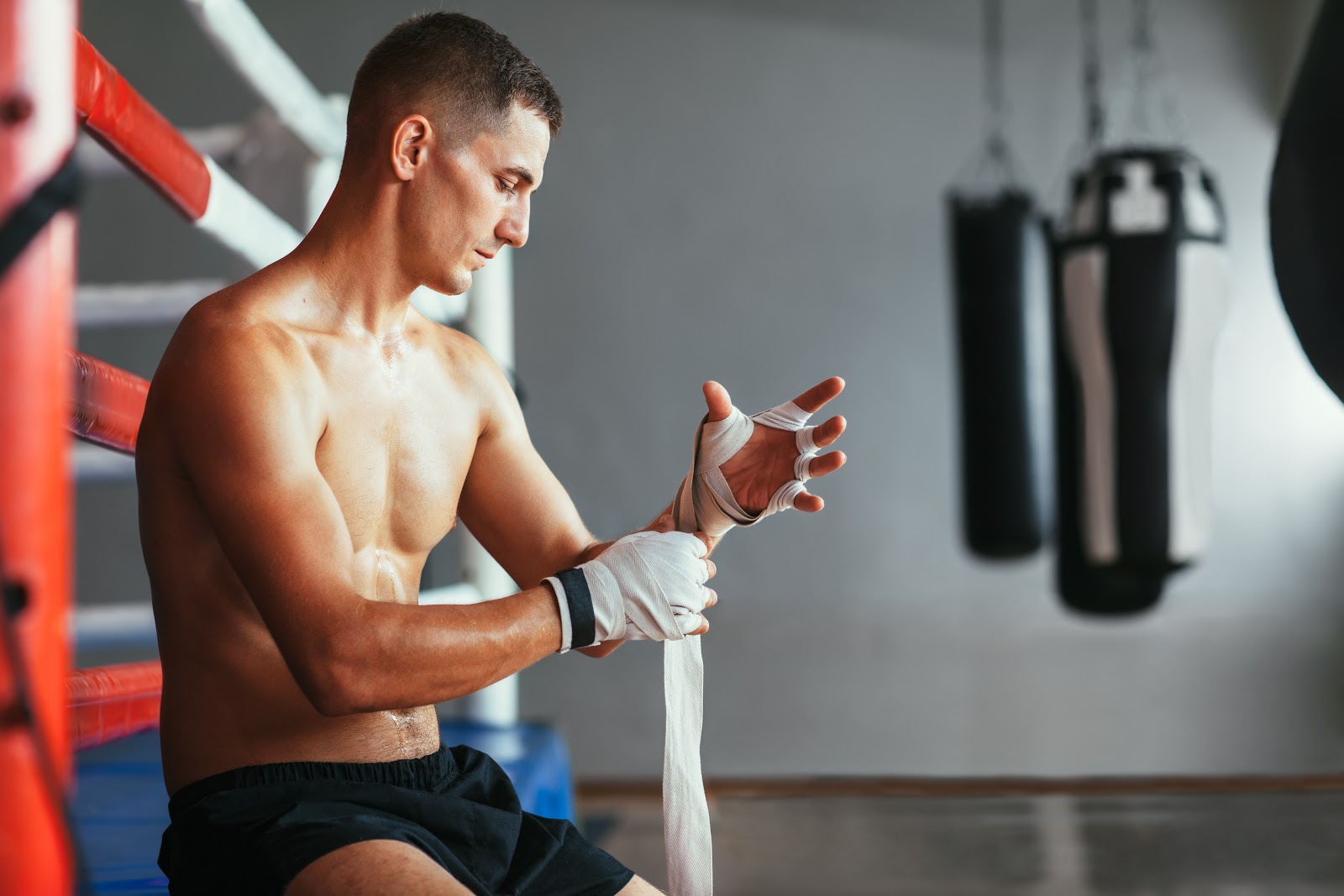Shirtless man wrapping his hands sitting on the edge of the boxing ring a boxing gym.